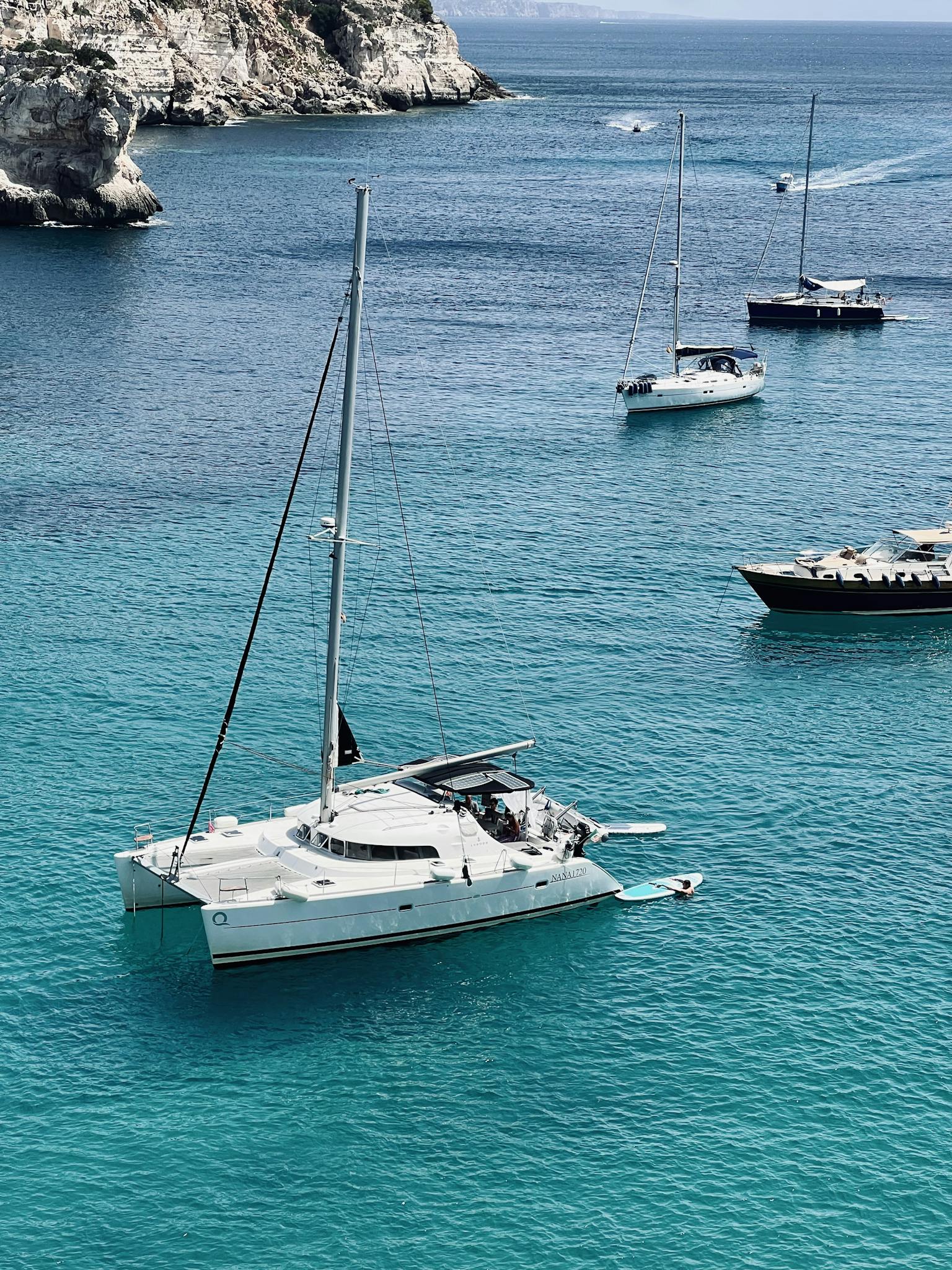 A serene view of a yacht and sailboats cruising in crystal clear turquoise waters near rocky cliffs.